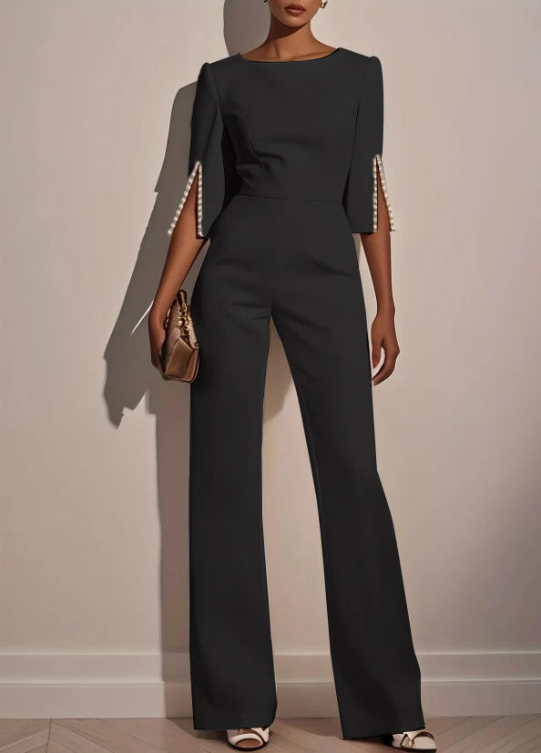 Woman in a black jumpsuit with pearl-trimmed sleeves holds a gold clutch in a minimalist indoor setting.