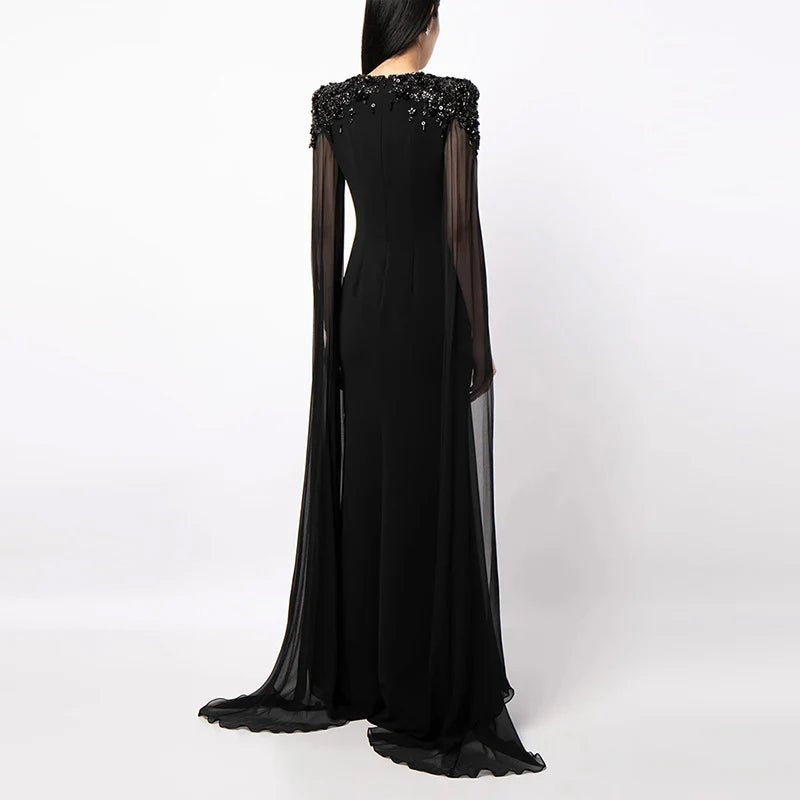 Back view of black dress showing intricate shoulder embellishments and flowing sheer cape in a studio.