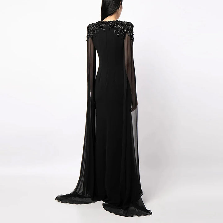 Back view of black dress showing intricate shoulder embellishments and flowing sheer cape in a studio.