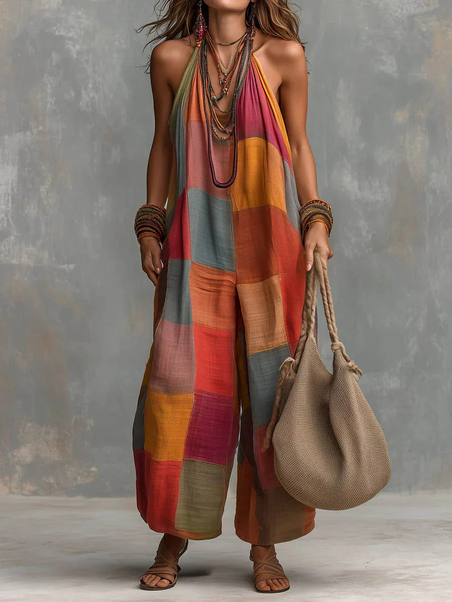 Bohemian jumpsuit in colorful patchwork, worn with layered necklaces and bangles. Model carries a woven bag, set against a textured gray backdrop.