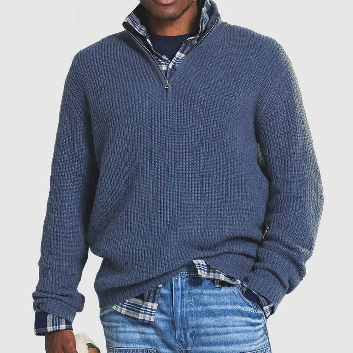 Man wearing a blue zip sweater over a plaid shirt with jeans, holding a hat in a studio setting.