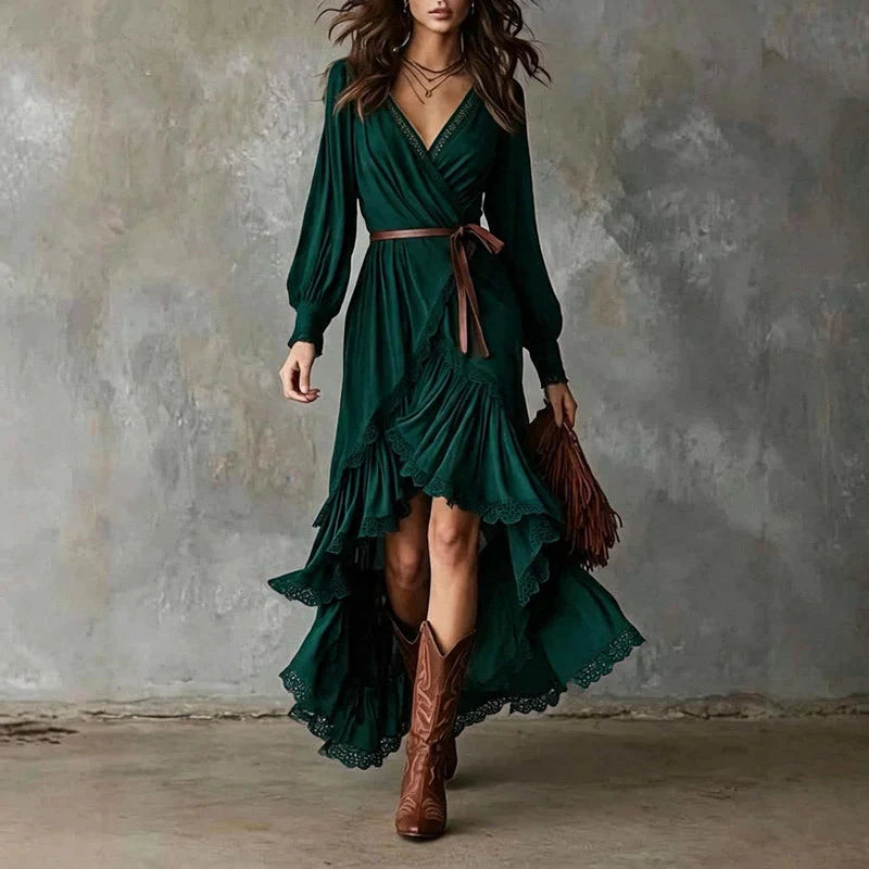 Woman in green wrap dress with long sleeves, lace trim, layered skirt. Holding brown fringe bag, wearing cowboy boots, gray backdrop.