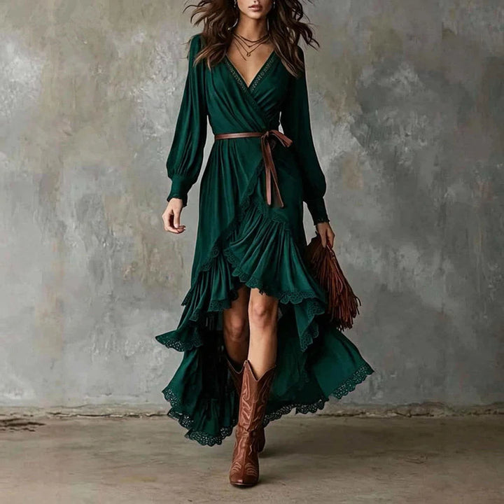 Woman in green wrap dress with long sleeves, lace trim, layered skirt. Holding brown fringe bag, wearing cowboy boots, gray backdrop.
