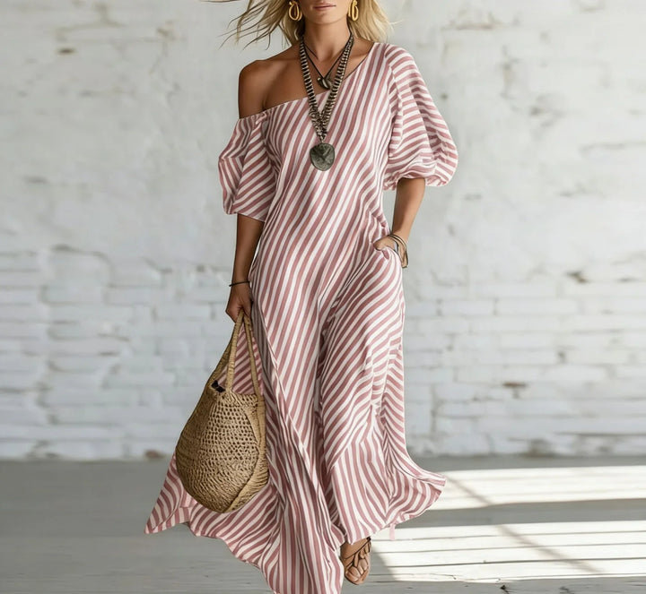 Woman wearing a pink and white striped off-shoulder maxi dress, accessorized with a necklace and earrings, holding a woven bag, in a bright, minimalist indoor setting.