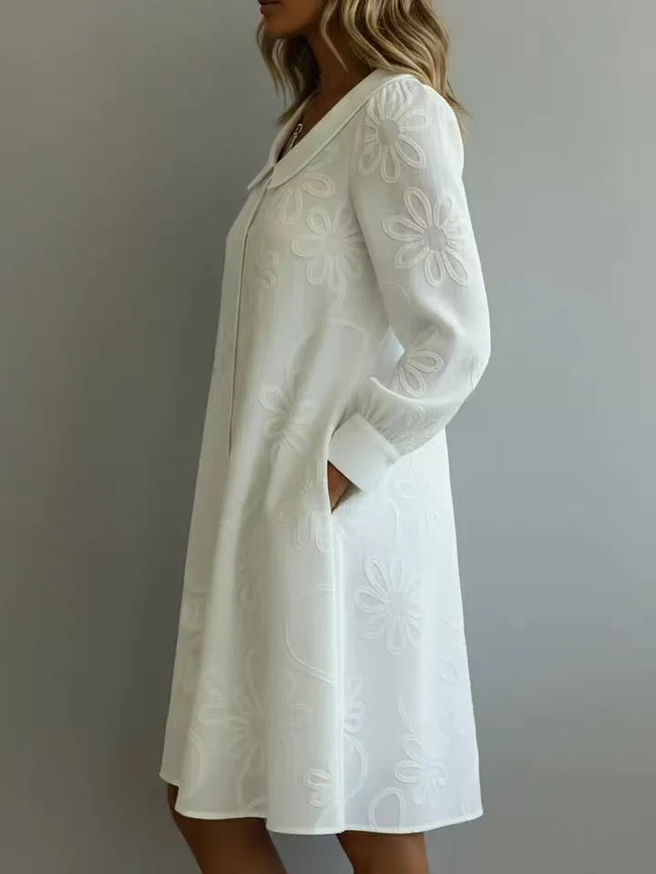 White midi dress with floral embroidery, collar, and long sleeves, shown in side profile in a neutral indoor setting.