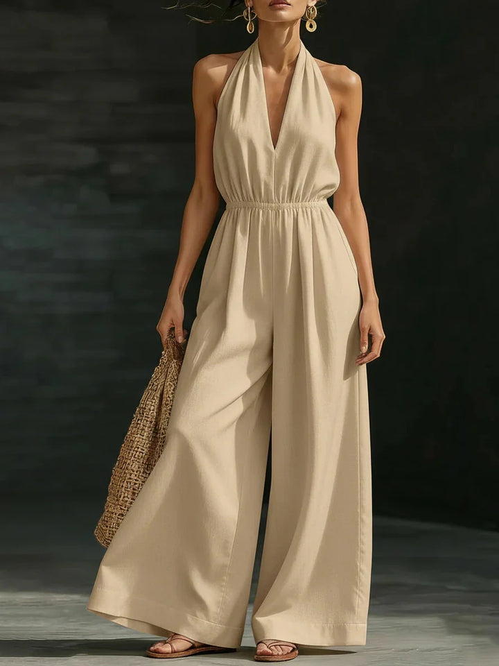 Woman in a beige jumpsuit featuring a halter neck and wide legs, paired with a woven bag, in a dark studio setting.