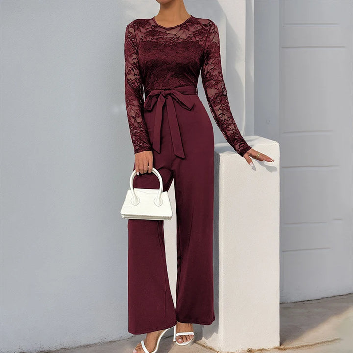 Chic burgundy jumpsuit with lace details and tie waist, carrying white handbag, posed outdoors against a light backdrop.