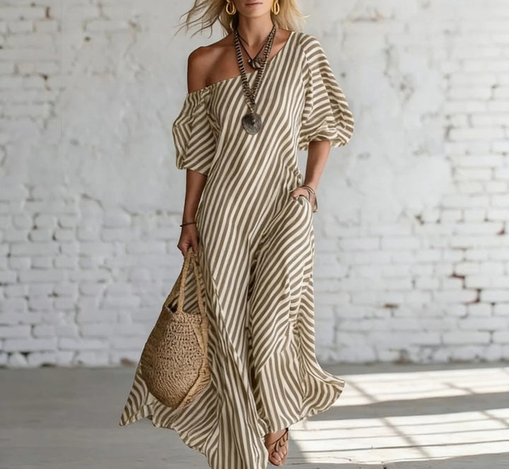 Woman wearing a beige and white striped off-shoulder maxi dress, accessorized with a necklace and earrings, holding a woven bag, in a bright, minimalist indoor setting.
