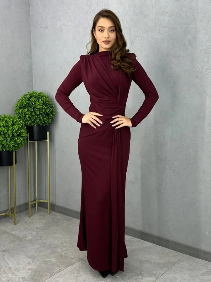 Model wearing burgundy bodycon maxi dress with long sleeves, hands on hips, in an indoor space with plants.