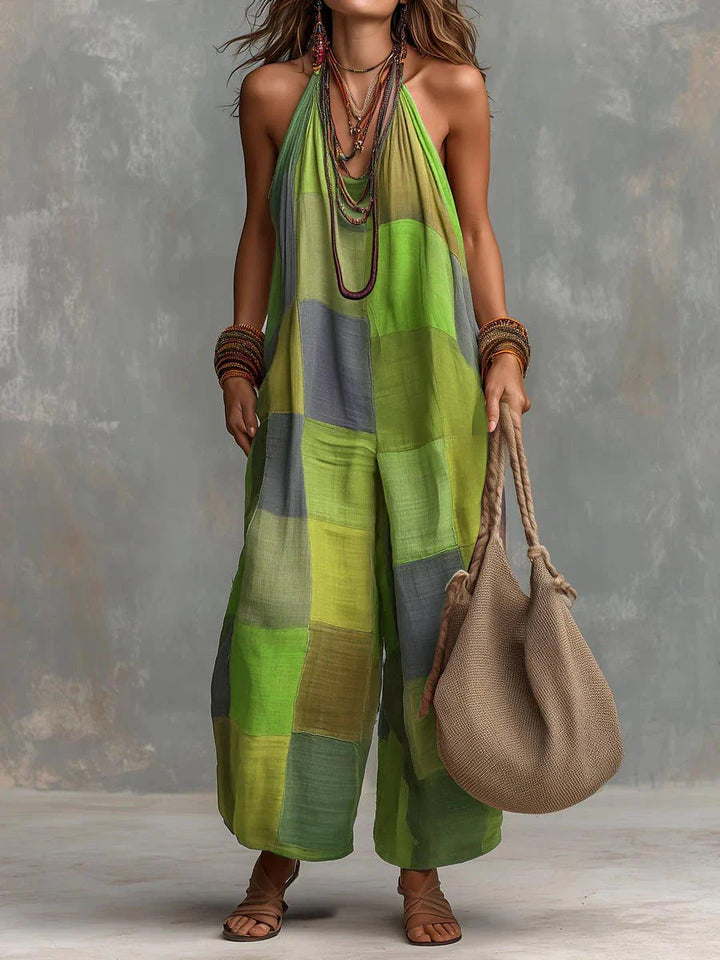 Green and gray patchwork bohemian jumpsuit, accessorized with necklaces and bangles. Model holds a woven bag, in front of a gray textured wall.