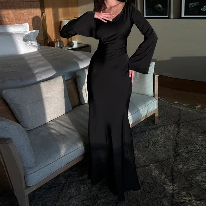 Black elegant maxi dress with long sleeves, modeled in a warmly lit bedroom with a plush bed and stylish decor.