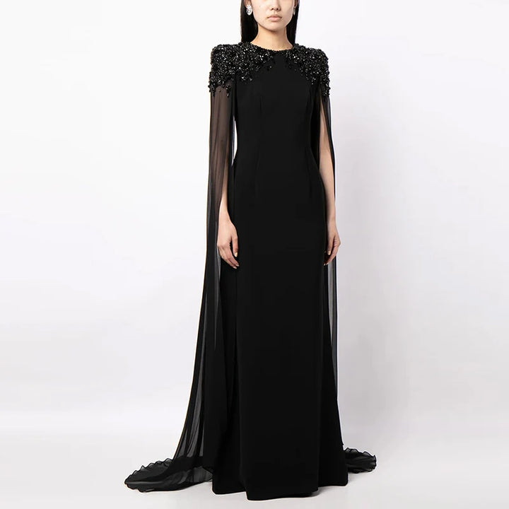 Black elegant dress with embellished shoulders and sheer cape sleeves, styled in a studio setting.