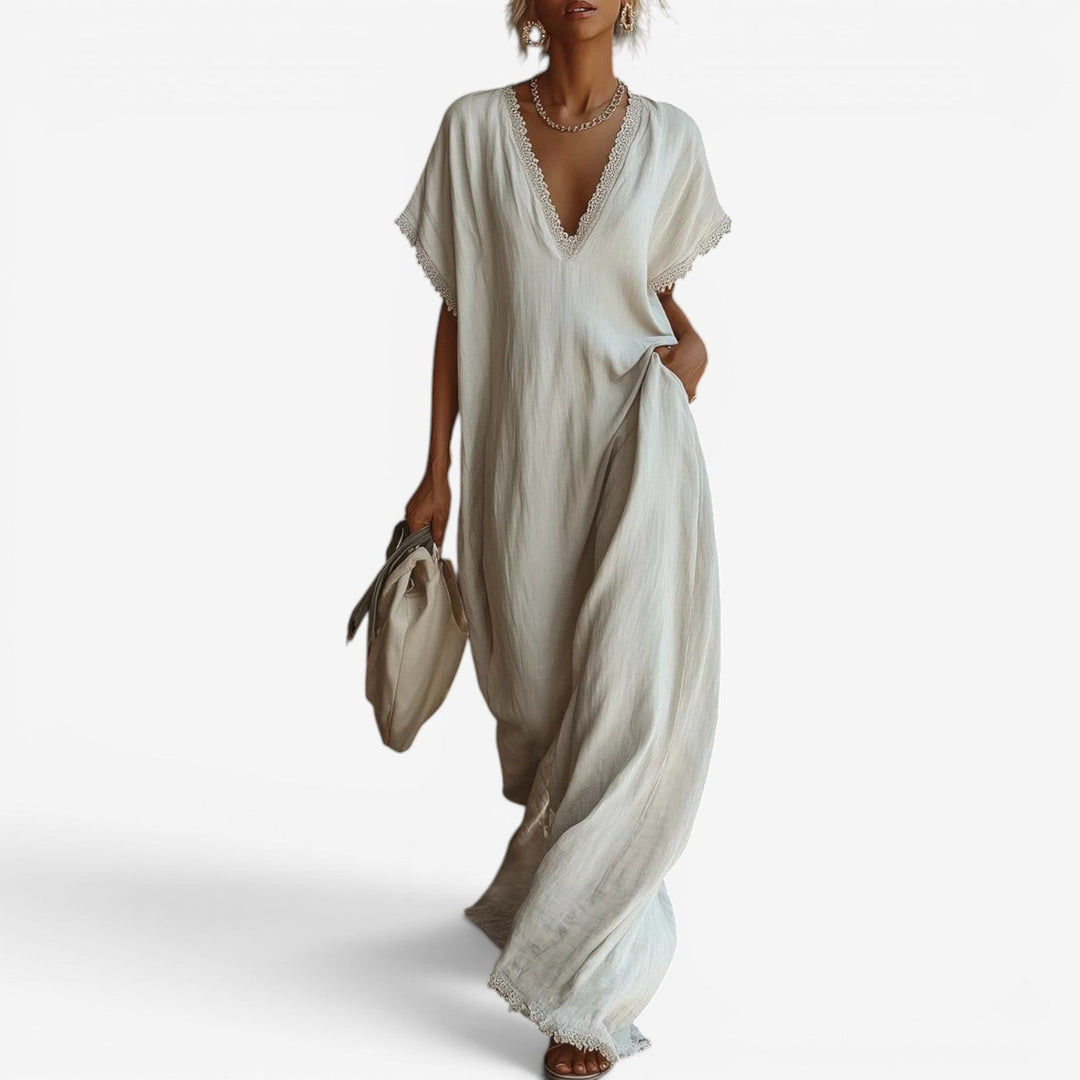 Flowing white maxi dress with lace-trimmed V-neckline, held by a woman carrying a beige bag against a plain background.