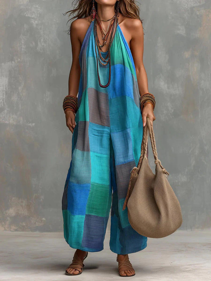 Blue and green patchwork bohemian jumpsuit, complemented by necklaces and bangles. Model with a woven bag, standing against a gray textured backdrop.