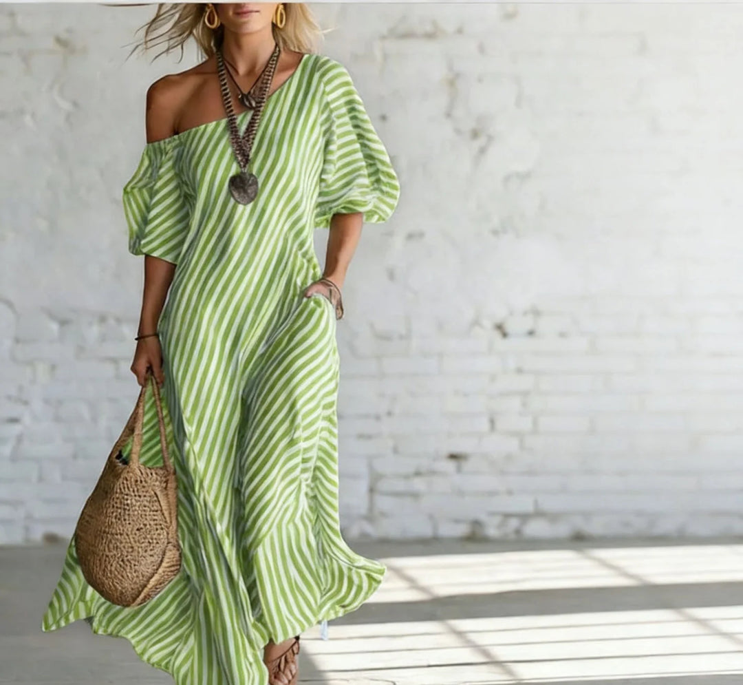 Woman wearing a green and white striped off-shoulder maxi dress, accessorized with a necklace and earrings, holding a woven bag, in a bright, minimalist indoor setting.