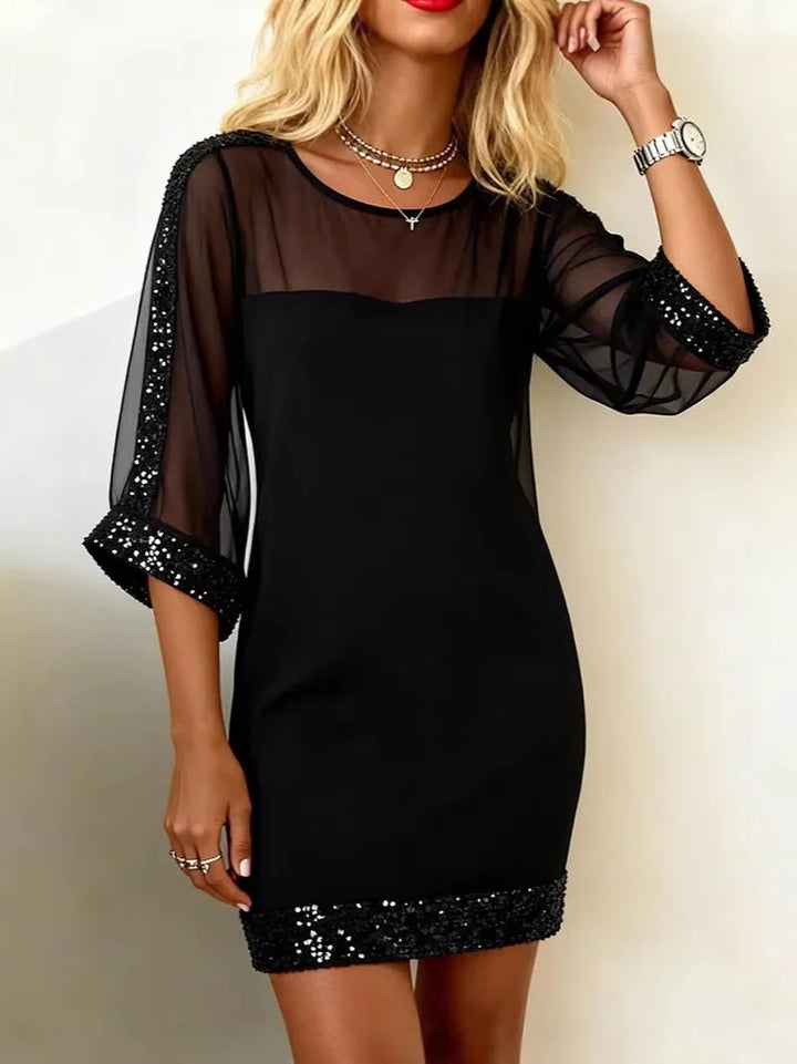Elegant black dress with sheer sleeves and sparkly trim, worn indoors against a light wall.