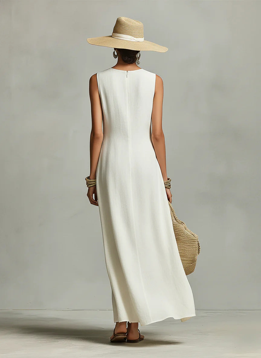 Back view of a woman in an ivory maxi dress and hat, holding a woven bag, in a studio setting.