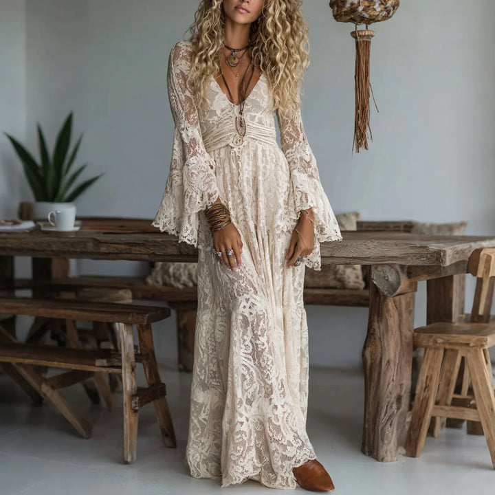 Woman in a flowing, cream maxi dress with lace details, standing in a rustic indoor setting with wooden furniture.