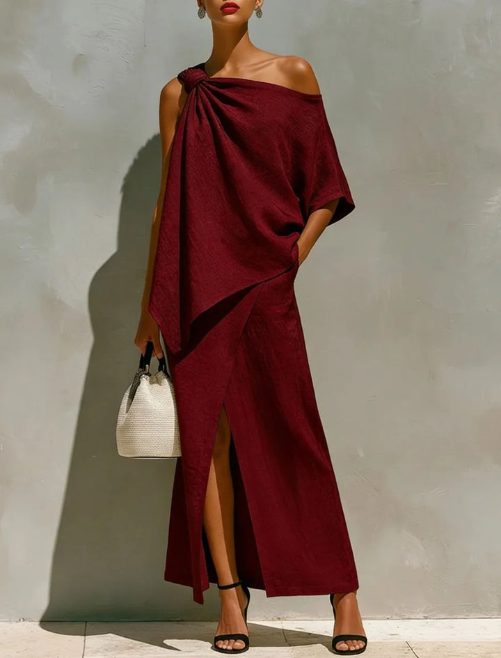 Elegant burgundy two-piece set with an off-shoulder top and long skirt with a slit, shown in a bright studio, styled with black heels and a woven bag.