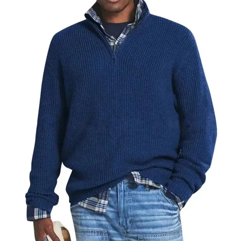 Man wearing a navy zip sweater over a plaid shirt with jeans, holding a hat in a studio setting.