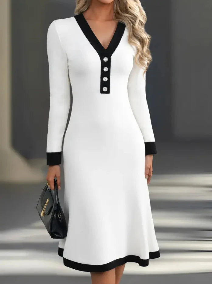 White long-sleeve dress with black trim and buttons, styled with a black handbag, set in a contemporary interior.
