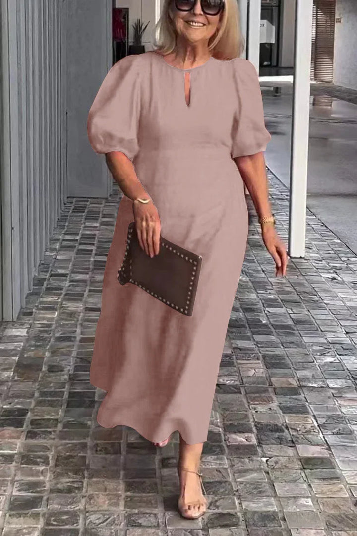 Woman in peach puff sleeve dress, holding clutch, walking on tiled path with modern gray wall backdrop.
