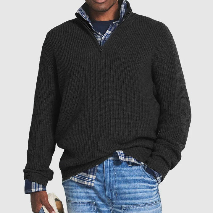 Man wearing a black zip sweater over a plaid shirt with jeans, holding a hat in a studio setting.