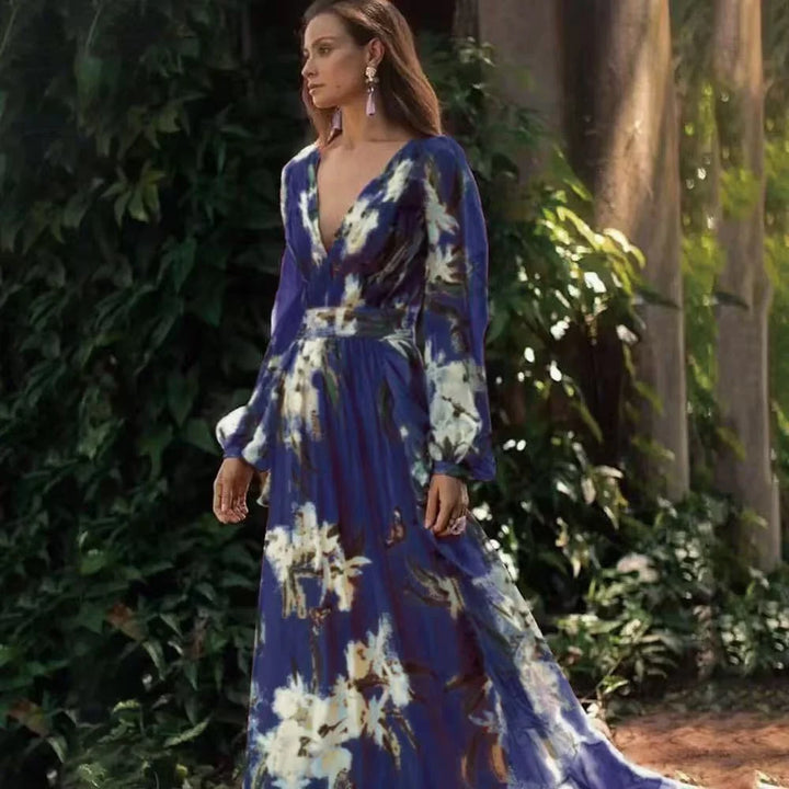 Woman in a flowing blue floral maxi dress with long sleeves, standing outdoors by lush greenery.
