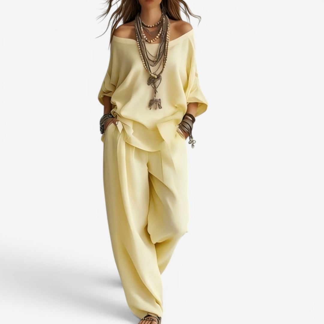 Elegant set in light yellow featuring off-shoulder top, flowing pants, and layered accessories. Studio background.