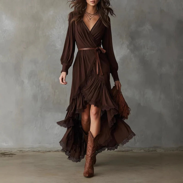 Woman in brown wrap dress with long sleeves, lace trim, layered skirt. Holding brown fringe bag, wearing cowboy boots, gray backdrop.