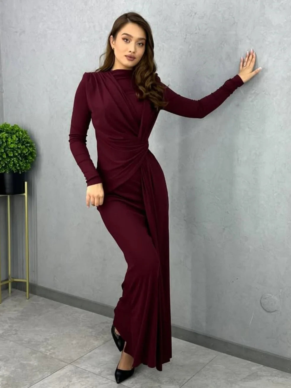 Person modeling a burgundy bodycon maxi dress with long sleeves, leaning against a wall indoors with a plant nearby.