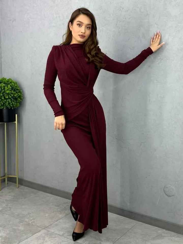Person modeling a burgundy bodycon maxi dress with long sleeves, leaning against a wall indoors with a plant nearby.