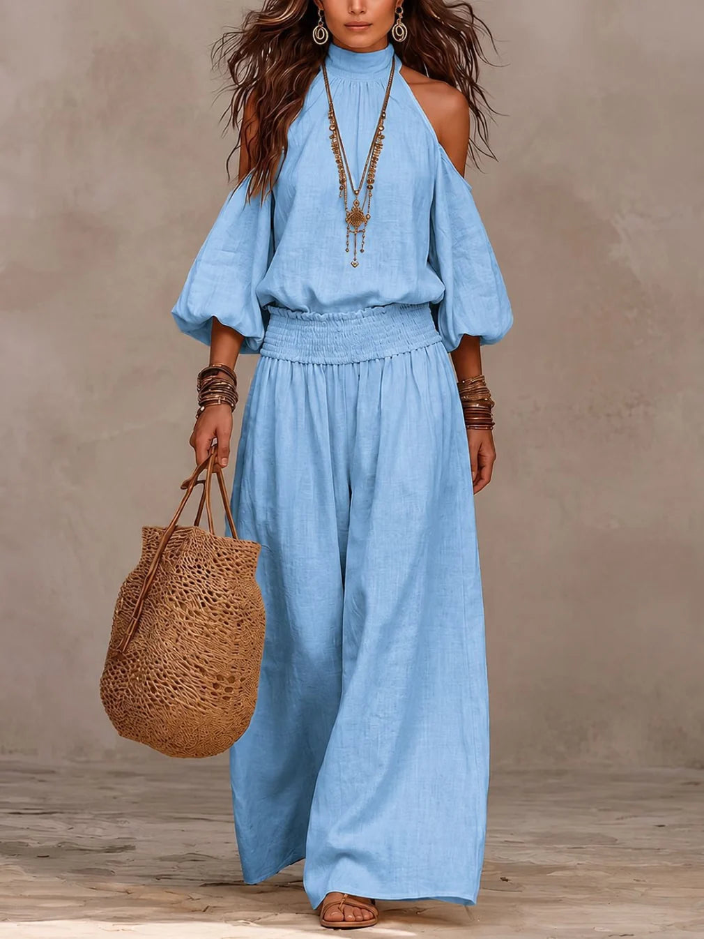 Model displays a light blue off-shoulder jumpsuit, accessorized with necklaces and a brown woven bag, in a simple studio setting.