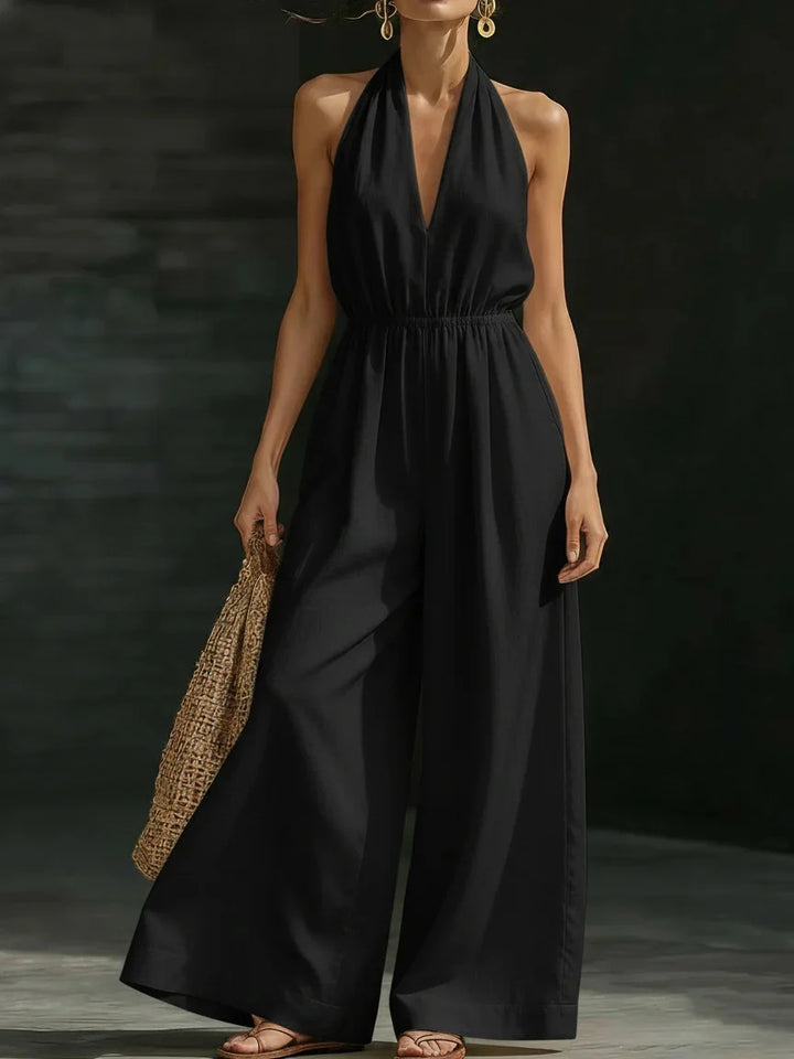 Woman in an elegant black jumpsuit with halter neck and wide legs, accessorized with a woven bag, in a dim studio.
