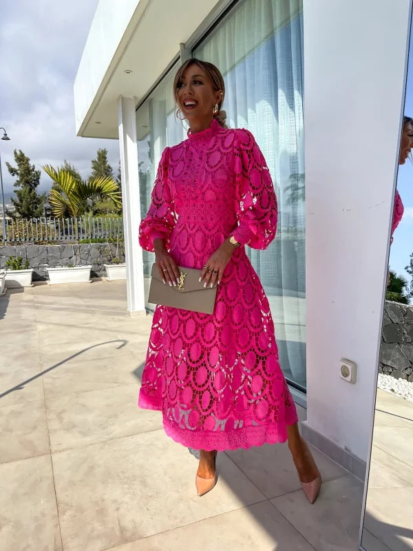 Pink lace dress in an outdoor setting, accessorized with a beige clutch and nude heels, posing by a window with greenery in view.