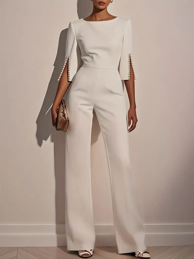 Woman in an ivory jumpsuit with pearl-accented sleeves carries a gold clutch in a minimalist indoor setting.