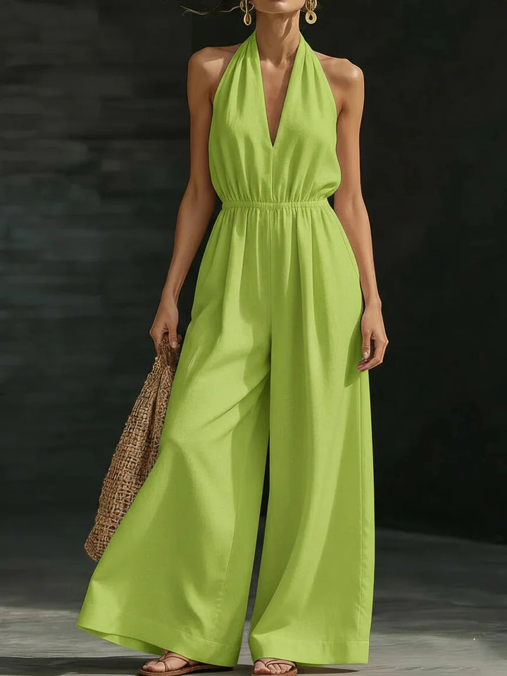 Woman in a bright green jumpsuit with halter neck and wide legs, holding a woven bag, set in a dark studio environment.