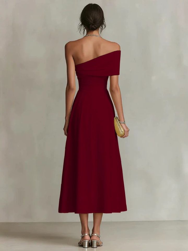 Back view of a woman in a burgundy one-shoulder maxi dress, holding a clutch, in a neutral studio setting.