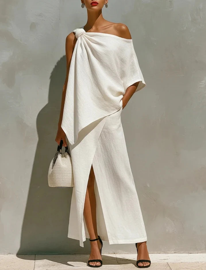 Sophisticated white two-piece set with an off-shoulder top and long skirt with a slit, featured in a light-filled studio, accessorized with black heels and a woven bag.