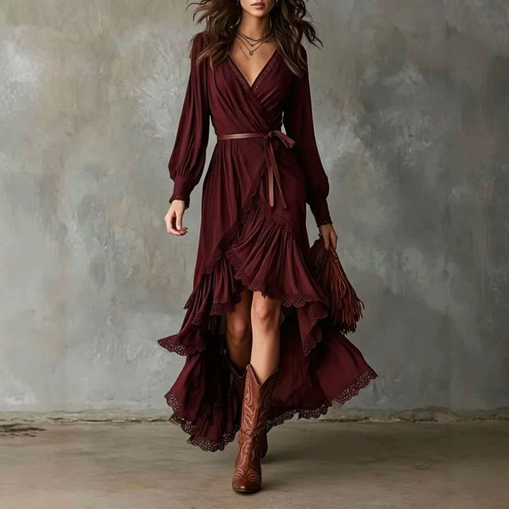 Woman in burgundy wrap dress with long sleeves, lace trim, layered skirt. Holding brown fringe bag, wearing cowboy boots, gray backdrop.
