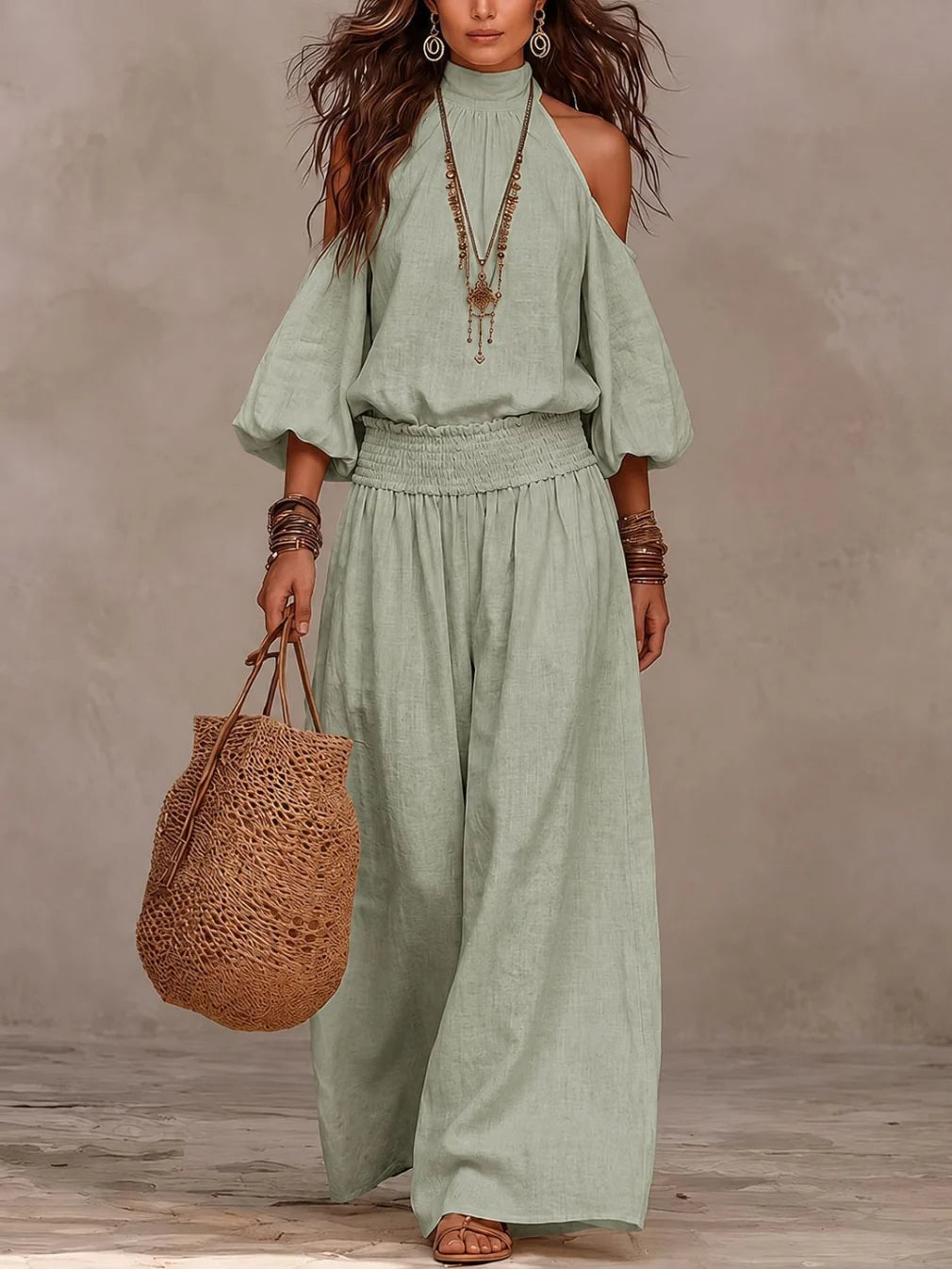 Model wearing sage green jumpsuit with off-shoulder design, accessorized with jewelry and holding a woven bag in a neutral setting.