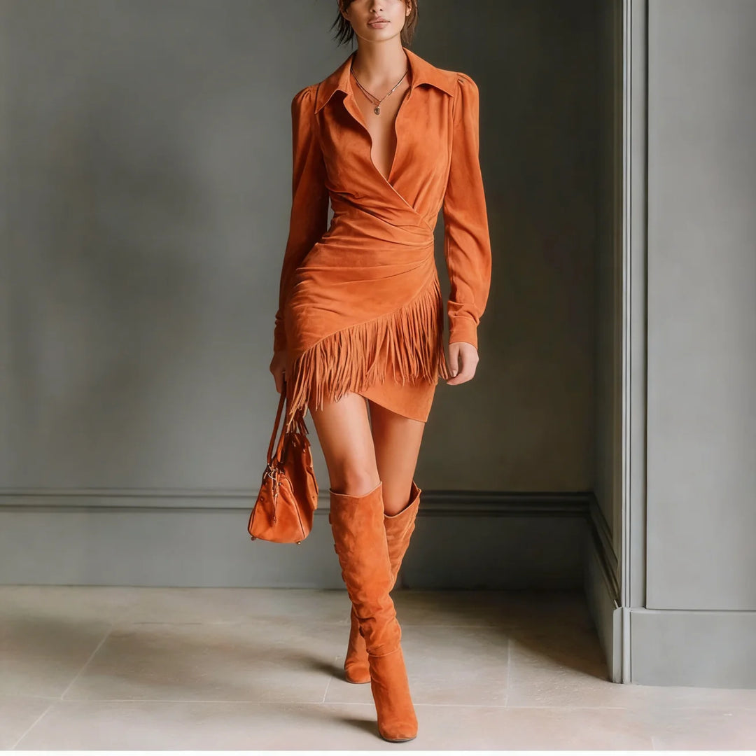 Fashionable woman in an orange mini dress with fringe and knee-high boots, posing indoors against a gray backdrop.