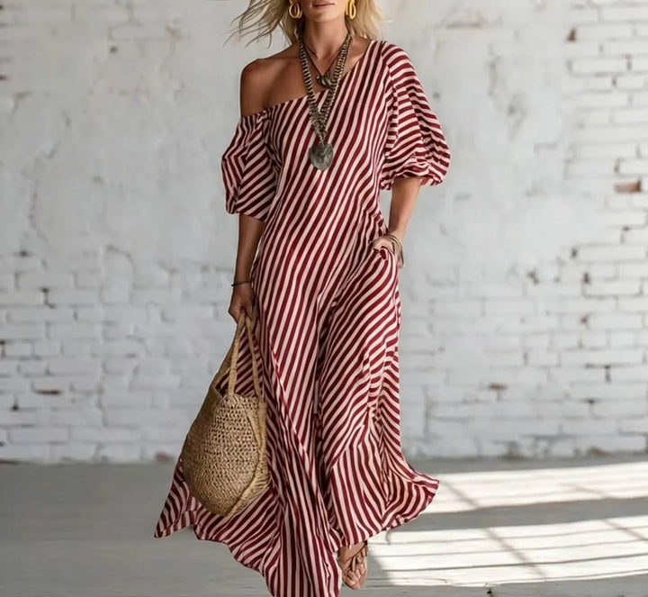 Woman wearing a red and white striped off-shoulder maxi dress, accessorized with a necklace and earrings, holding a woven bag, in a bright, minimalist indoor setting.