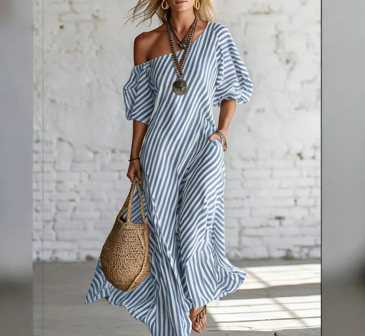 Woman wearing a blue and white striped off-shoulder maxi dress, accessorized with a necklace and earrings, holding a woven bag, in a bright, minimalist indoor setting.