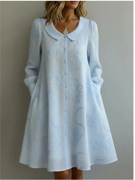 Soft blue midi dress with floral embroidery, collar, and long sleeves in a neutral indoor setting.