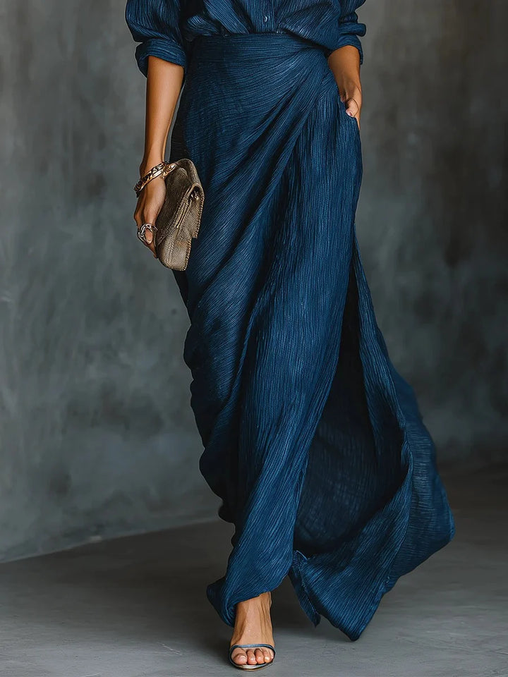 Lower view of a blue maxi dress showcasing its flowing skirt, paired with strappy sandals and a clutch in an indoor setting.