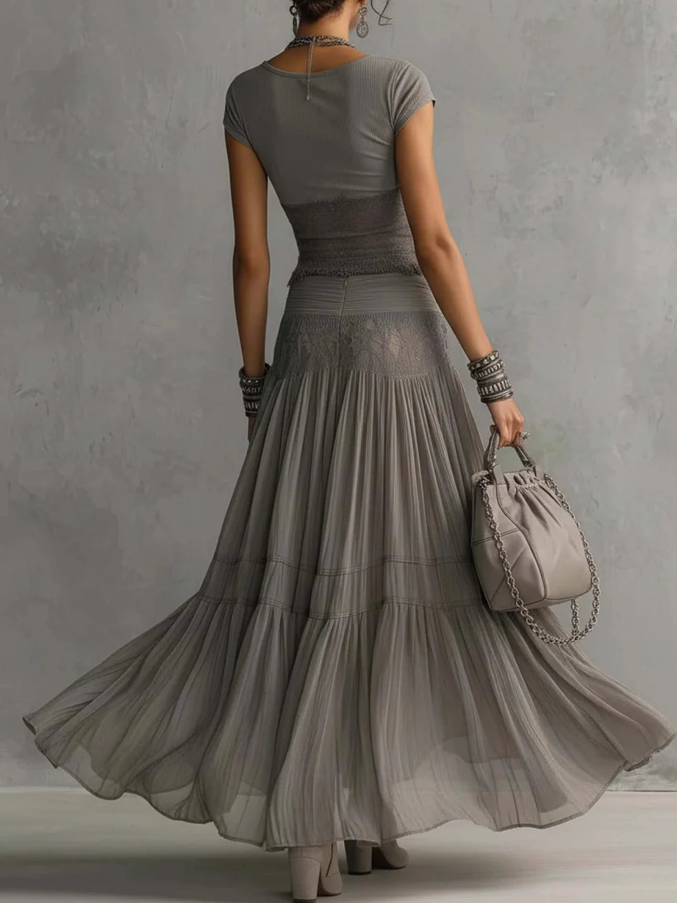 Back view of a woman in a flowing gray maxi skirt set, holding a small bag with chain straps in a studio setting.