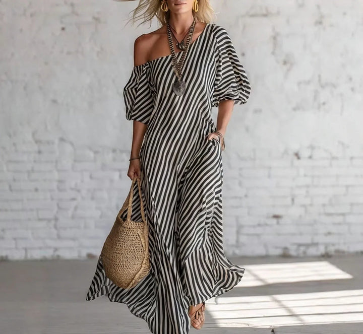 Woman wearing a black and white striped off-shoulder maxi dress, accessorized with a necklace and earrings, holding a woven bag, in a bright, minimalist indoor setting.