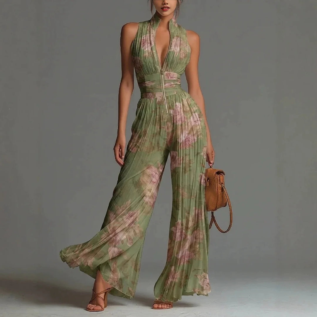 Model wearing elegant green floral jumpsuit with pleated legs, standing in studio holding a brown bag.