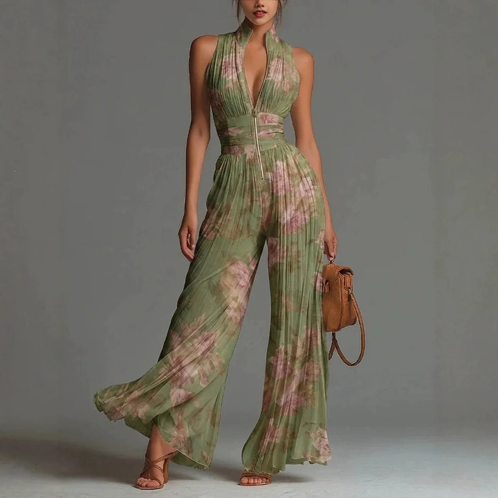 Model wearing elegant green floral jumpsuit with pleated legs, standing in studio holding a brown bag.